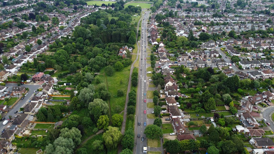 An aerial view of a suburban residential street in Friern Barnet, showing houses with gardens on both sides of a straight road, with multiple cars parked along the kerb. On the left side of the road, there is a strip of greenery and trees, forming a small park or green space with pathways winding through the trees. The image captures the scene during daytime with natural lighting, illustrating the calm, organized layout typical of a neighbourhood suitable for house removals and home relocation services, such as those offered by Man with Van Friern Barnet. This view highlights the area's accessibility for moving activities, including the loading process involving furniture transport and packing within a typical residential environment.