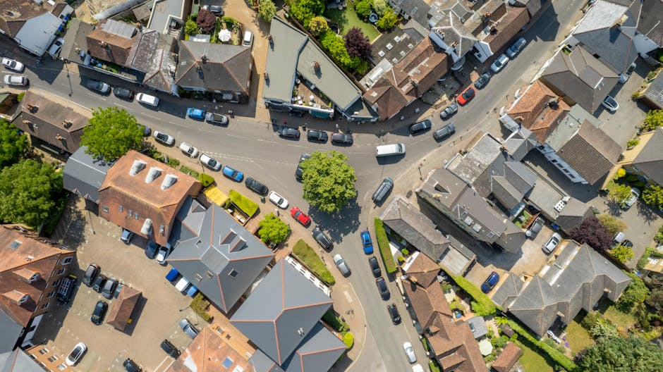 An aerial view of a suburban residential street in Friern Barnet, showing houses with gardens on both sides of a straight road, with multiple cars parked along the kerb. On the left side of the road, there is a strip of greenery and trees, forming a small park or green space with pathways winding through the trees. The image captures the scene during daytime with natural lighting, illustrating the calm, organized layout typical of a neighbourhood suitable for house removals and home relocation services, such as those offered by Man with Van Friern Barnet. This view highlights the area's accessibility for moving activities, including the loading process involving furniture transport and packing within a typical residential environment.