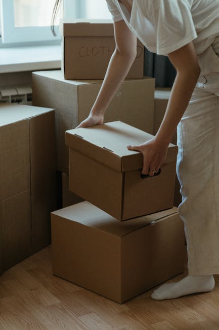 A person dressed in light clothing is seen moving a cardboard box during a home relocation, with several other boxes stacked nearby inside a well-lit room with large windows. The individual appears to be carefully lifting or placing the box, which is labeled 'CLOTHES' in red marker on the top. The boxes are made of brown corrugated cardboard, with some covered in plastic wrap for protection. They are positioned on a wooden floor, close to a door or window frame, indicating an indoor environment prepared for furniture transport and packing during a house removal. Visible furniture includes a cardboard box and possibly a larger item wrapped for moving. The scene captures the typical loading process where household items are being organized for transportation with the assistance of a professional service like Man with Van Friern Barnet, specializing in removals for small flats in Friern Barnet.