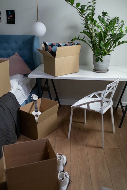 Inside a residential room during a home relocation process, a white desk is positioned against a light-colored wall, with two cardboard moving boxes placed on and near it. One open box on the desk contains padding and packing materials, while another cardboard box on the floor holds an electric guitar, with additional empty boxes nearby. A white modern chair with a geometric backrest is situated next to the desk. The background features a bed with a blue upholstered headboard, pillows, and a hanging spherical lamp beside the bed. To the right, a large potted plant with broad green leaves adds a natural element to the space. The wooden flooring suggests a domestic environment, and the scene reflects stages of packing and furniture transport in a house move, with clear visibility of packing materials, furniture, and placement within the room, ideal for a removals company's visual content supporting house relocation services.