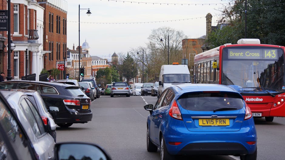 A busy street scene showing parked cars along both sides of the road, including a white van and a blue hatchback vehicle, with some cars parked partially on the pavement. An orange-red Metroline bus w